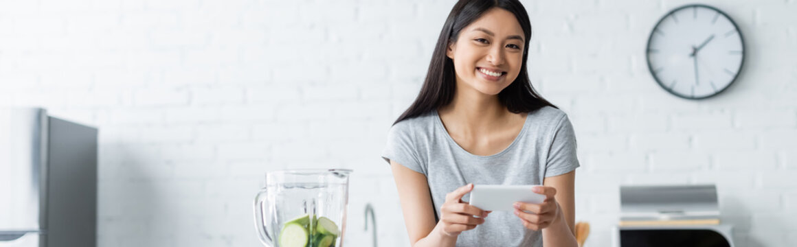 cheerful asian woman taking photo on smartphone near blender with sliced zucchini, banner.