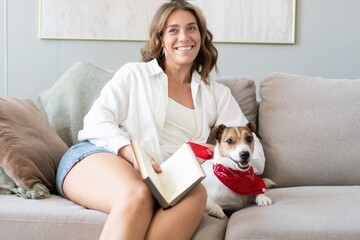Young woman reading book and her cute dog on sofa at home. Lovely pet.