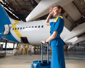 Beautiful air stewardesses in bright blue uniform looking away while standing with suitcase in front of passenger aircraft. Occupation concept