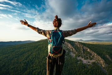 A traveler girl with a backpack is standing on the edge of the mountain, a rear view. A young woman with a backpack standing on the edge of a cliff and looking at the sky with her hands raised.
