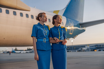 Two beautiful air hostesses in blue uniform smiling away, standing in front of a big passenger...