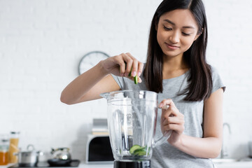 smiling asian woman adding fresh zucchini into electric shaker in kitchen.