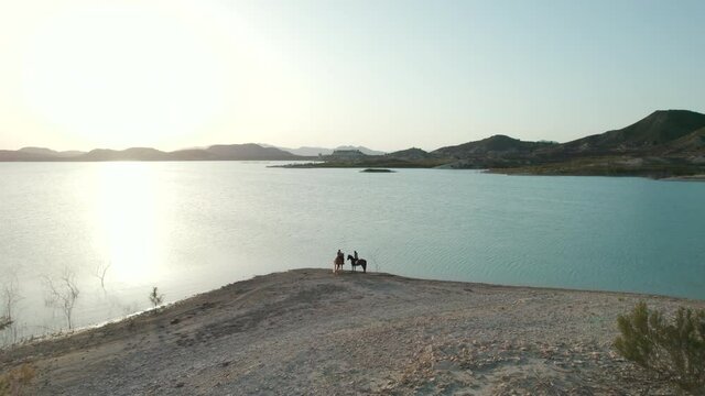 Aerial drone point view two young adult caucasian women rides on horses outdoor on the coast of Embalse de La Pedrera lake, Orihuela. Spain. Concept of hobby, equestrian sport and active lifestyle