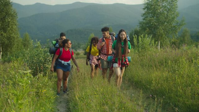 Group of cheerful attractive diverse multiracial travelers with backpacks communicating, enjoying active lifestyle and summer nature, ascending mountain hill while hiking in mountain during vacations.