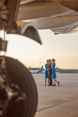 Full length shot of male pilot posing for photoshoot together with two air stewardesses in blue uniform, standing in an airport terminal at sunset. Aircraft, aircrew concept