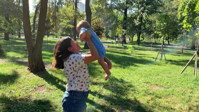 a brunette mother of European appearance holds her laughing baby boy in her arms, whirls with him, lifts up and kisses on a sunny day in a green park