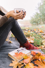 Unrecognizable woman taking picnic on lake shore. Slim girl drinking coffee or tea from white polka dot cup. On plaid lying book, glasses and colorful maple leaves. Warming beverage for autumn season.