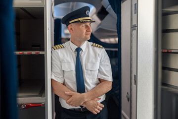 Thoughtful male pilot in uniform and hat keeping arms crossed and looking aside while standing inside of the airplane. Transportation, aircrew concept © Yaroslav Astakhov