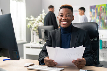 A young man's first day at work after graduation, a dark-skinned smiling boy sits behind a desk in an office room, holding documents, reports, getting acquainted with the functioning of company