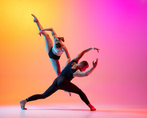 Two dancers, stylish sportive couple, male and female models dancing contemporary dance on colorful gradient yellow pink background in neon light.