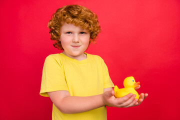 Photo of funny candid ginger little boy hold rubber duck toy wear yellow t-shirt isolated red color...