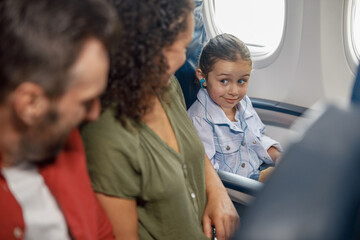 Fototapeta na wymiar Cute little girl wearing earphones while sitting on the plane, listening to music, looking at her parents, traveling together with family. Travel, vacation concept