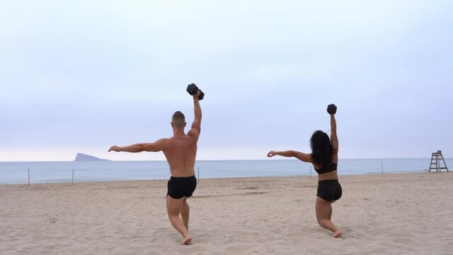 Diverse strong sportsman and sportswoman doing exercises with dumbbells on beach at