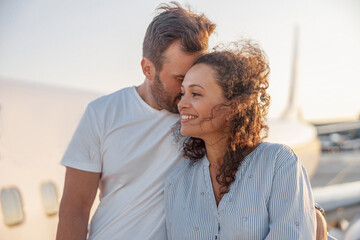 Portrait of lovely couple of tourists, man and woman looking happy while standing outdoors ready for boarding the plane at sunset. Vacation, lifestyle, traveling concept