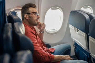 Handsome caucasian man in casual wear and glasses looking away, sitting on the plane near the window. Relax, travel, vacation, transportation concept - Powered by Adobe