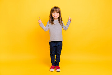 Full body photo of cute little blond hair girl point empty space wear shirt jeans sneakers isolated on yellow backgound