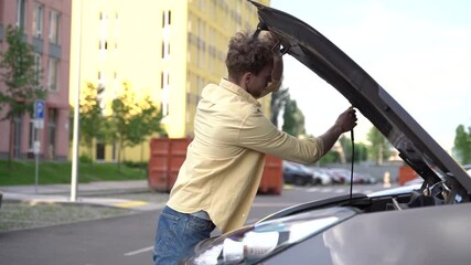 Guy in the middle of the street opened the hood of his car. A young man inspects the car engine