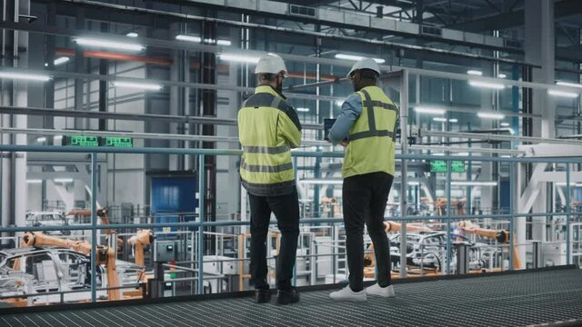 Two Car Factory Specialists in High Visibility Vests Using Laptop Computer. Engineers Discussing Automotive Industrial Manufacturing Facility Process of Vehicle Production. Automated Assembly Plant.