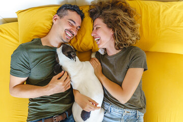 Happy moment of couple with pet at home on bed. Horizontal top view playing with pet indoors.