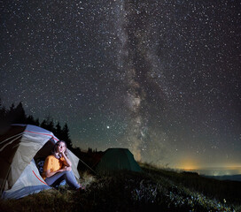 Fantastic view of night starry sky over grassy hill with camp tents and female traveler. Charming...