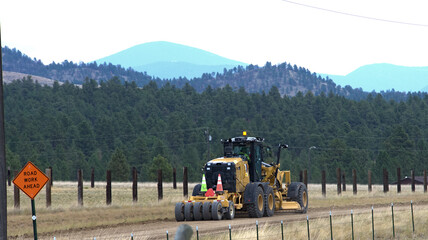 Road grader on rural road with fencing, trees and mountains in the background.