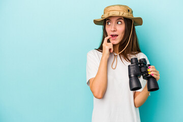 Young caucasian explorer woman holding binoculars isolated on blue background relaxed thinking about something looking at a copy space.