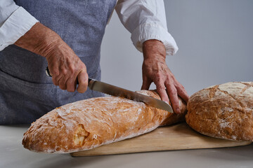 The hands of an elderly person hold and cut bread with a knife. Freshly baked bread in senior wrinkled hands. Selective focus