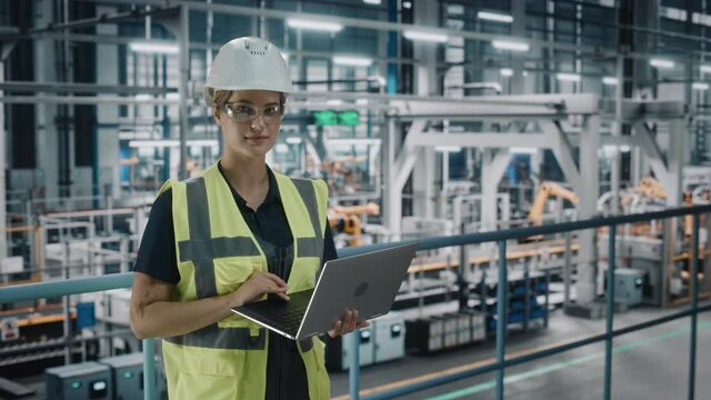 Portrait of Female Automotive Industry 4.0 Engineer in Safety Uniform Using Laptop at Car Factory Facility. Happy Assembly Plant Specialist Working on Manufacturing Modern Electric Vehicles.