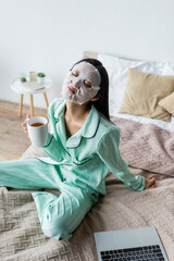 asian woman in sheet mask sitting on bed with cup of tea and closed eyes near laptop.