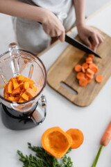 partial view of blurred woman cutting carrot near blender with pumpkin.