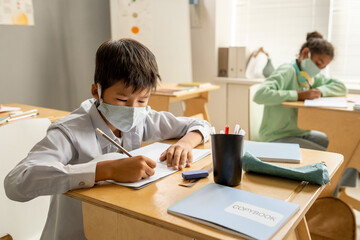 Obraz premium Diligent Asian schoolboy in protective mask writing in copybook against classmate