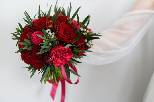 The bride holds a beautiful bouquet of red roses and peonies at arm's length.