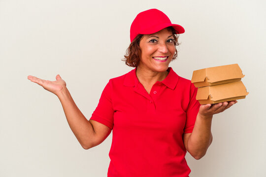Middle age delivery woman taking burguers isolated on white background showing a copy space on a palm and holding another hand on waist.