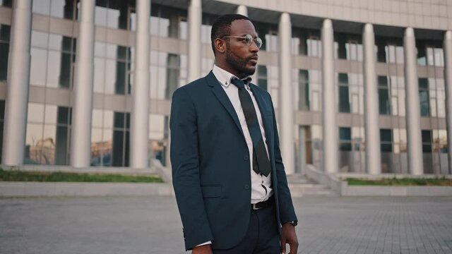 Zoom in portrait of confident african american employee wearing eyeglasses standing with bag outdoors, tracking shot