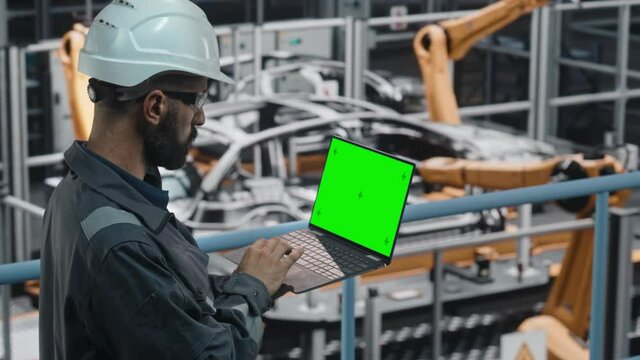 Car Factory Engineer in Work Uniform Using Laptop Computer with Green Screen Mockup Display. Working with Software at Automotive Industrial Manufacturing Facility Dedicated for Vehicle Production.
