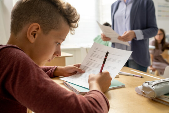 Youthful schoolboy carrying out test by desk against teacher and classmates