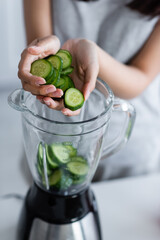 cropped view of blurred woman adding cut cucumber into electric shaker.
