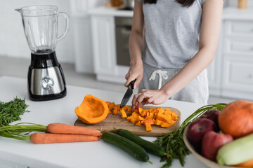 cropped view of woman cutting ripe pumpkin near fresh vegetables and electric blender.