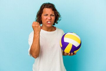 Middle age latin woman playing volleyball isolated on blue background showing fist to camera, aggressive facial expression.