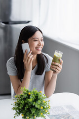 joyful asian woman with glass of fresh smoothie talking on mobile phone near blurred plant.