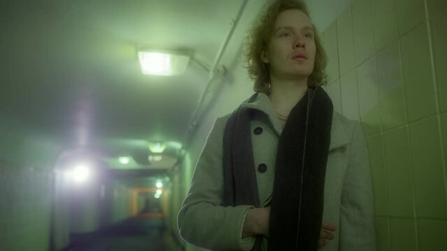 An attractive Caucasian man blond with long hair in a coat stands in the underpass and throws a scarf around his neck.