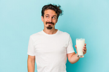 Young caucasian man holding a glass of milk isolated on blue background dreaming of achieving goals...