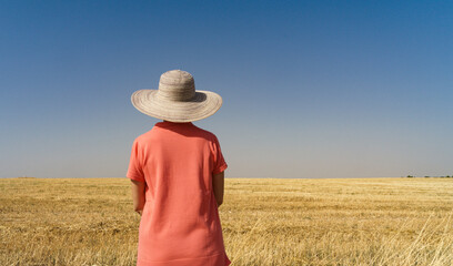 Mature woman with her back turned and wearing a hat with her back to the camera and in front of a landscape of cereal crops already mowed with blue sky.