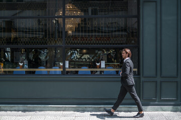 Fototapeta premium Successful stylish man in formal black suit walking on the street near dark-green wall. High quality photo