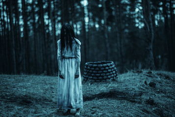 Girl in image of scary zombie walks in dark forest near stone well. - obrazy, fototapety, plakaty