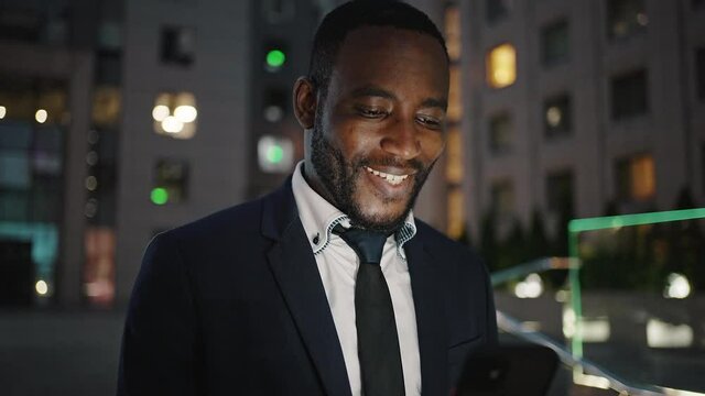Good news. Happy african american businessman reading great message on cellphone and smiling, standing in evening city