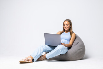 Beautiful girl is sitting on bean bag couch and is looking laptop isolated on white background.