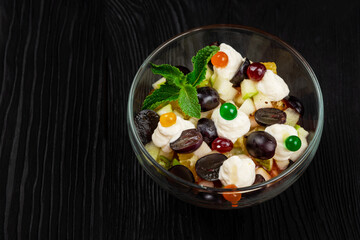 Salad from grapes apples pears kiwi oranges with mascarpone chease and cream. Healthy fresh fruit summer salad in glass bowl on black wooden background with copy space.