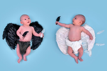 A newborn baby boy with white angel wings and a child with black evil wings, blue studio background