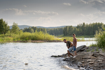 Fototapeta na wymiar Young man and teenage boy enjoying time by lake and throwing small stones into water
